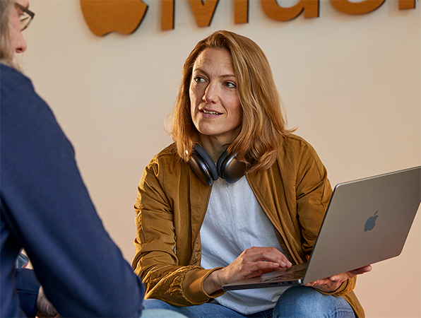 Dos personas del equipo de Desarrollo de Empresas y Ventas de Apple conversando, una de ellas con una MacBook en la mano y sentada frente a una pared con el logotipo de Apple Music.