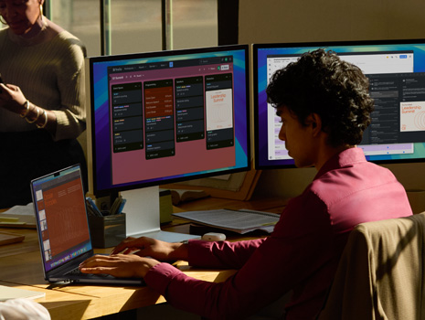 A woman sitting at a table in a high-rise building. Behind her is a large window revealing other buildings. In front of her is an open MacBook.